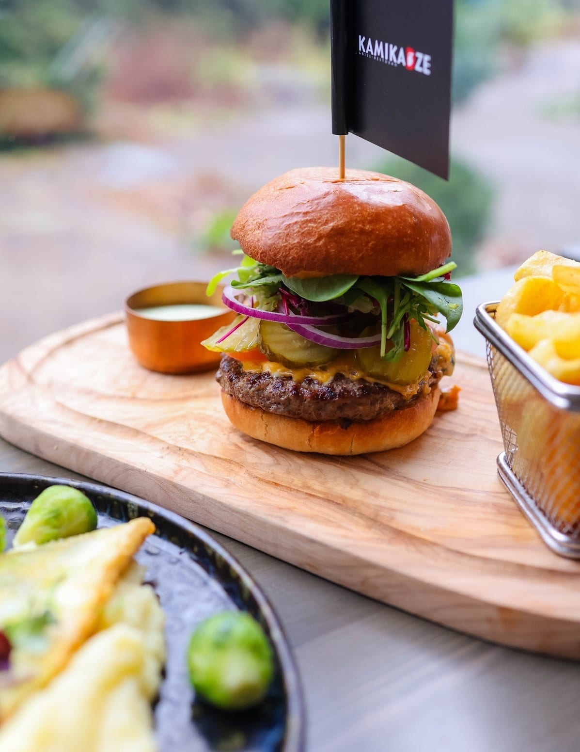 Burger mit Salat und Gurken auf einem Holzbrett, daneben Pommes im Körbchen